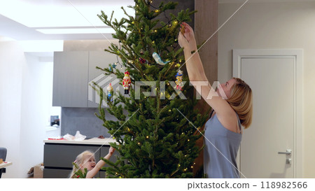 Young mother and her daughter are adorning a tall christmas tree in their contemporary apartment with bright lights and colorful decorations. New Year's Eve with family 118982566