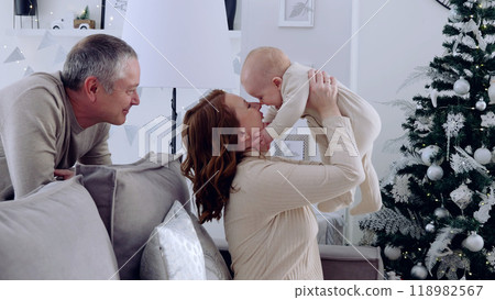 Young family enjoys holiday moments, playing with their baby beside the christmas tree in the living room. A day with family before Christmas. New Year's Eve with family 118982567