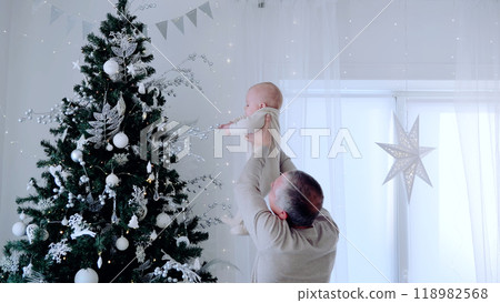 Grandfather lifting his baby grandson to touch the top of a decorated christmas tree, creating a heartwarming holiday moment. A day with family before Christmas. New Year's Eve with family 118982568