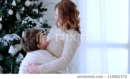 Mother and son share a joyful holiday embrace by their beautifully decorated christmas tree, creating a heartwarming and magical scene. A day with family before Christmas. New Year's Eve with family 118982579
