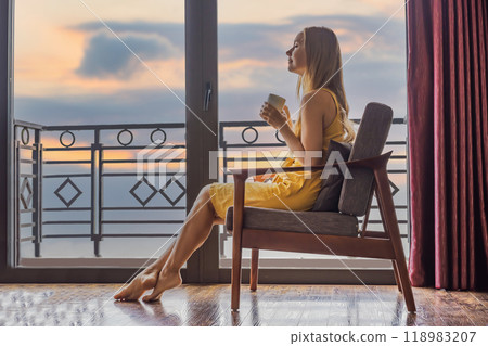 A young woman holding coffee cup while sittinhg on chair on balcony , looking at mountains and green nature. Woman in Sapa in the fog, Northwest Vietnam. Vietnam travel concept. UNESCO heritage 118983207