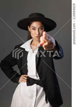 woman in black suit and wide-brimmed hat imitates shooting from a pistol directly into the camera. on gray background, vertical photo 118984029
