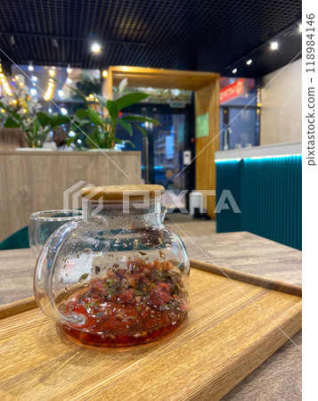 Glass teapot with an herbal tea sits on a wooden tray in a modern cafe. Ambient lighting and contemporary decor create a warm and inviting atmosphere Glass teapot with an herbal tea sits on a wooden tray in a modern cafe. Ambient lighting and contemporary decor create a warm and inviting atmosphere 118984146
