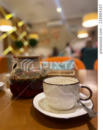 Delicate teacup alongside a clear teapot with blooming herbal tea captures a cozy tea time in a stylish, modern cafe. Inviting ambiance is highlighted by warm lighting and contemporary decor. 118984167
