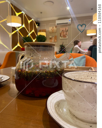 Delicate teacup alongside a clear teapot with blooming herbal tea captures a cozy tea time in a stylish, modern cafe. Inviting ambiance is highlighted by warm lighting and contemporary decor. 118984168
