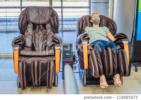 Beautiful young woman wearing a medical mask due to the coronavirus COVID 19 relaxing on the massage chair in airport or in the mall Beautiful young woman wearing a medical mask due to the coronavirus COVID 19 relaxing on the massage chair in airport or in the mall 118985873