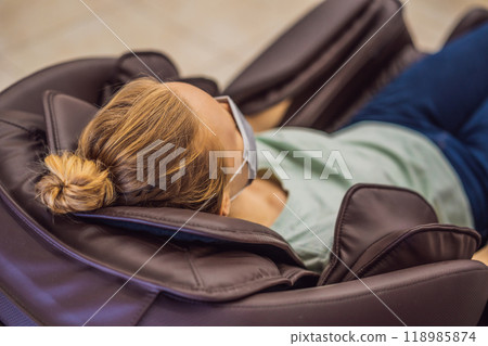 Beautiful young woman wearing a medical mask due to the coronavirus COVID 19 relaxing on the massage chair in airport or in the mall Beautiful young woman wearing a medical mask due to the coronavirus COVID 19 relaxing on the massage chair in airport or in the mall 118985874