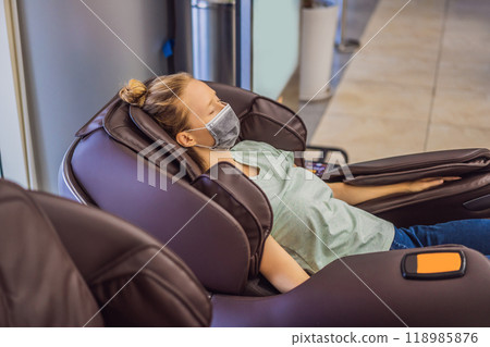 Beautiful young woman wearing a medical mask due to the coronavirus COVID 19 relaxing on the massage chair in airport or in the mall Beautiful young woman wearing a medical mask due to the coronavirus COVID 19 relaxing on the massage chair in airport or in the mall 118985876