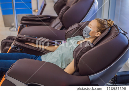 Beautiful young woman wearing a medical mask due to the coronavirus COVID 19 relaxing on the massage chair in airport or in the mall Beautiful young woman wearing a medical mask due to the coronavirus COVID 19 relaxing on the massage chair in airport or in the mall 118985896