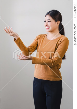 Cheerful asian woman smiles while gesturing with open hands presenting or explaining something. Cheerful asian woman smiles while gesturing with open hands presenting or explaining something. 118986958