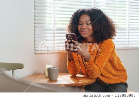 Happy young african american woman in orange sweater using smartphone while sitting in a cozy room. Happy young african american woman in orange sweater using smartphone while sitting in a cozy room. 118986993