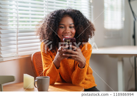 Happy young african american woman in orange sweater using smartphone while sitting in a cozy room. Happy young african american woman in orange sweater using smartphone while sitting in a cozy room. 118986994