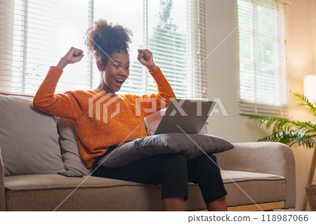 Young black woman celebrating success while working on laptop in cozy living room. 118987066