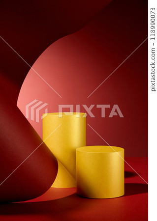 A minimalist product display features golden cylinder podiums against vibrant red paper, creating a dynamic and eye-catching composition with a curved and flowing form. 118990073