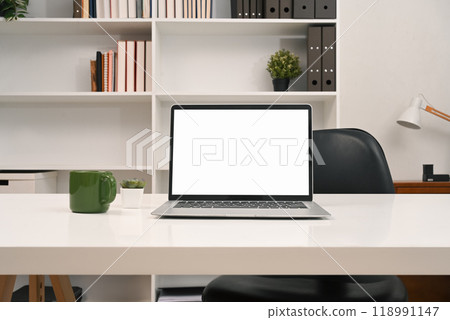 A laptop computer with empty display and office supplies on a desk in front of a bookshelf 118991147