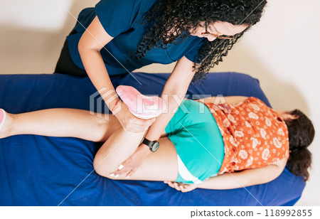 Top view of Chiropractor stretching knee to lying female patient, Knee flexion physiotherapy to female patient. Physiotherapist woman assisting knee to lying patient Top view of Chiropractor stretching knee to lying female patient, Knee flexion physiotherapy to female patient. Physiotherapist woman assisting knee to lying patient 118992855