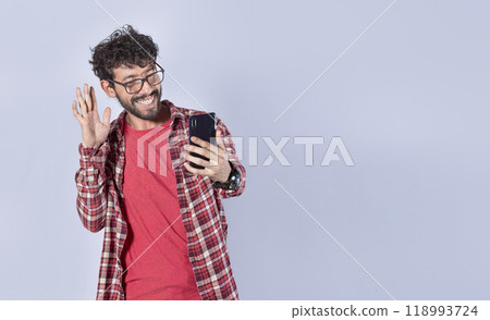 Smiling man making video call with his cellphone isolated, Positive teen guy making video call waving hands, Concept of smiling man saying hello by video call on cellphone Smiling man making video call with his cellphone isolated, Positive teen guy making video call waving hands, Concept of smiling man saying hello by video call on cellphone 118993724
