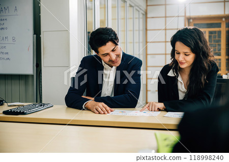 In an office setting, two colleagues engage in a meeting, focusing on teamwork, business strategy, and financial success. Their smiles and collaboration are evident. 118998240
