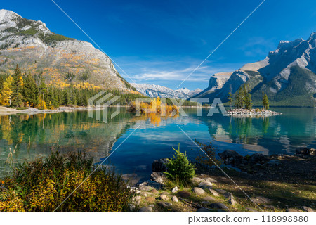Banff National Park Lake Minnewanka autumn landscape. Alberta, Canada. Majestic mountains, forests in yellow green color against blue sky reflected on the water. Beautiful natural scenery. 118998880