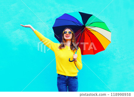 Happy smiling young woman holding colorful umbrella in autumn looking up on colorful blue background Happy smiling young woman holding colorful umbrella in autumn looking up on colorful blue background 118998938