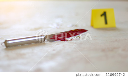 A bloodstained stainless steel knife lying on a concrete floor, with a blurred yellow number 1 sign nearby and blood splatter surrounding the scene, evoking a sense of crime investigation. 118999742