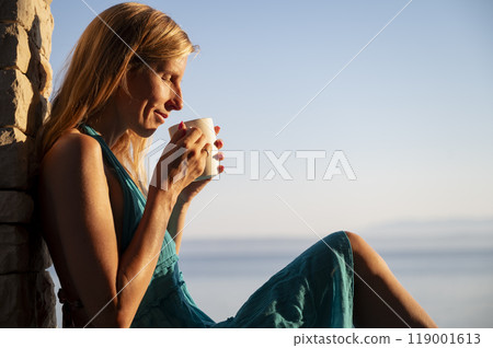 Young woman enjoying the smell of her coffee while she relaxes leaning on a stone wall by the sea 119001613