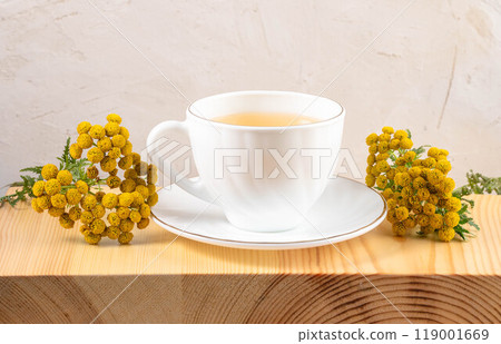 White cup on saucer of healthy herbal tansy tea and tansy flowers on textured wooden board close up. White cup on saucer of healthy herbal tansy tea and tansy flowers on textured wooden board close up. 119001669