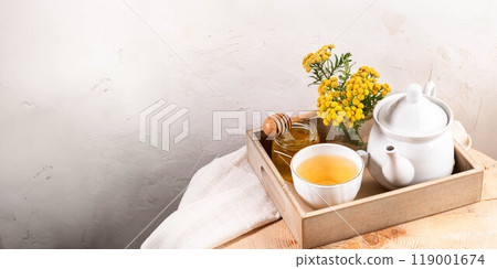 Tansy tea in teapot and cup, tansy flowers, honey in wooden tray on textured white wall backdrop. Tansy tea in teapot and cup, tansy flowers, honey in wooden tray on textured white wall backdrop. 119001674