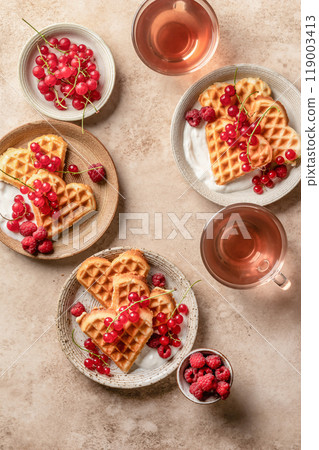 Homemade waffles with summer berries and yogurt served for sweet breakfast with tea on beige background. Heart shaped waffles top view Homemade waffles with summer berries and yogurt served for sweet breakfast with tea on beige background. Heart shaped waffles top view 119003413