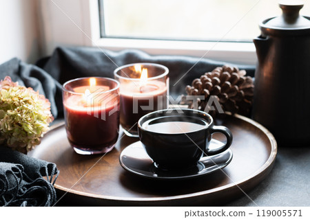 Cup of coffee, aroma candles served in wooden tray on windowsill. Winter holiday. Cozy home decor. Close up. Cup of coffee, aroma candles served in wooden tray on windowsill. Winter holiday. Cozy home decor. Close up. 119005571