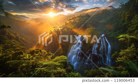 Sunrise over tranquil mountains illuminates a waterfall amidst lush greenery, creating a peaceful scene. 119006453