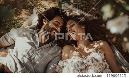 A couple lies on a blanket under a tree, laughing and pointing at cloud shapes as leaves rustle gently. 119006815