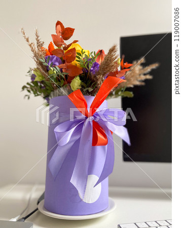 Colorful bouquet of flowers in a lavender box standing on a desk, tied with a vibrant red and purple ribbon. Bright, mixed arrangement adds a touch of nature and warmth to modern workspace 119007086