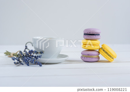 Delicious violet and yellow macarons and cup of latte or americano and branch of fragrant lavender on white wooden background. 119008221