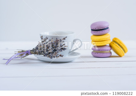 Delicious violet and yellow macarons and cup of latte or americano and branch of fragrant lavender on white wooden background. Delicious violet and yellow macarons and cup of latte or americano and branch of fragrant lavender on white wooden background. 119008222