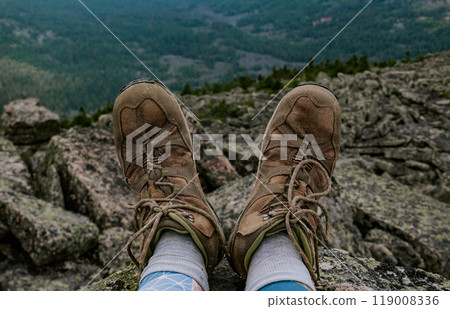 Feet in old brown trekking boots against a background of rocky and forested terrain. traveler shoe details 119008336