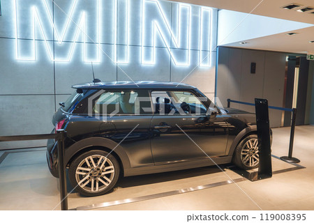 A pristine black Mini Cooper showcased indoors with a large illuminated MINI sign in a high-end dealership or automotive exhibition in London, UK. A pristine black Mini Cooper showcased indoors with a large illuminated MINI sign in a high-end dealership or automotive exhibition in London, UK. 119008395