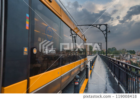 A yellow metro train travels on the iconic Dom Luis I Bridge in Porto, Portugal. The city's rooftops and buildings are visible under a dramatic overcast sky. A yellow metro train travels on the iconic Dom Luis I Bridge in Porto, Portugal. The city's rooftops and buildings are visible under a dramatic overcast sky. 119008418