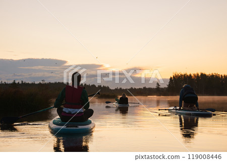 A serene scene of three individuals on stand-up paddleboards during sunrise on a tranquil lake. The sky glows with soft hues of orange and yellow, surrounded by nature. A serene scene of three individuals on stand-up paddleboards during sunrise on a tranquil lake. The sky glows with soft hues of orange and yellow, surrounded by nature. 119008446