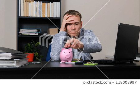 Man at an office table places coins into a pink piggy bank, deep in thought with organized paperwork and a laptop nearby 119008997