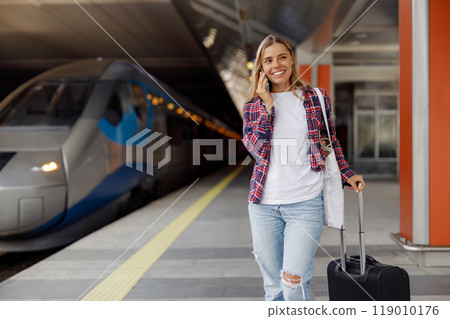 A cheerful traveler is at a bustling train station, happily waiting with their luggage in hand 119010176