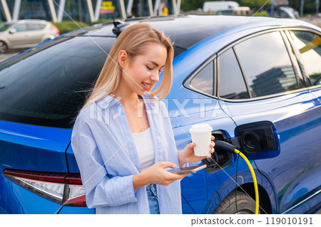 Woman charges her electric car while enjoying coffee and checking her smartphone in a parking area 119010191