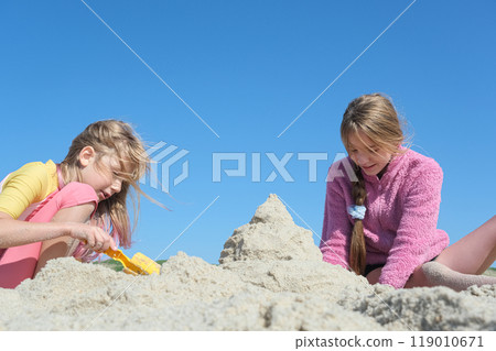 Two girls building a sandcastle on the beach under a clear blue sky. Two happy sisters having fun on the Atlantic ocean deserted beach. 119010671