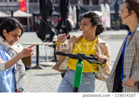 Three friends enjoy a fun ride on an electric scooter, laughing together in the vibrant city square 119014287