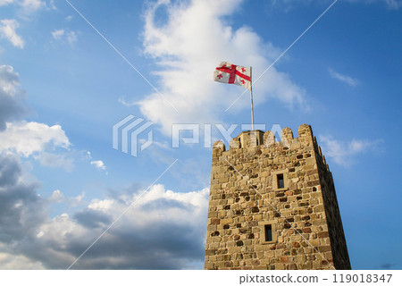 National flag of Georgia on the top of old stone tower flies waving against the background of a blue sky with clouds 119018347
