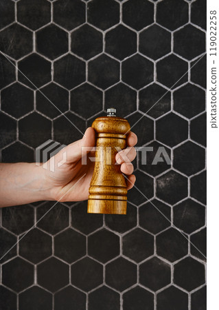 A hand holding a wooden pepper mill against a textured black hexagonal tile background 119022258