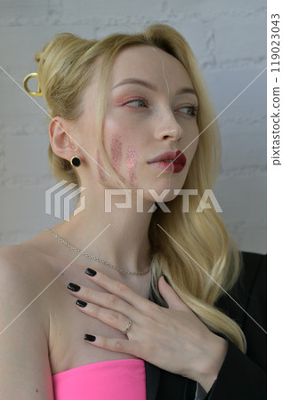 Close-up portrait of a blonde girl with different make-up on half of her face and a thoughtful look with hand on the chest on white background 119023043