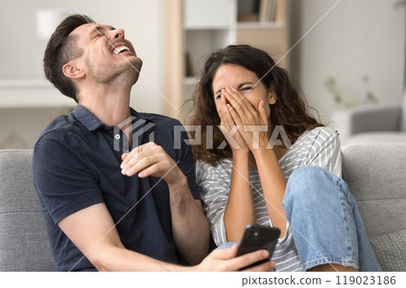 Joyful millennial couple holding smartphone on home sofa 119023186