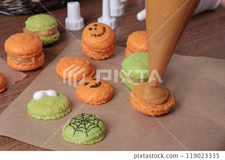 Process of making Halloween macarons. Anonymous squeezing caramel cream from pastry bag onto dough in kitchen. Baking macarons at home close-up. 119023335