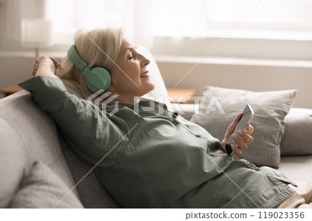 Elderly woman wearing wireless headphones, holding smartphone, enjoying music 119023356